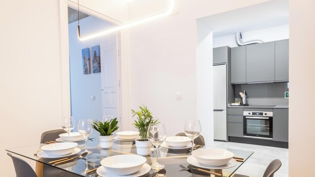 A modern dining area with a glass table, white plates, and wine glasses, leading to a kitchen with grey cabinets and a built-in oven.