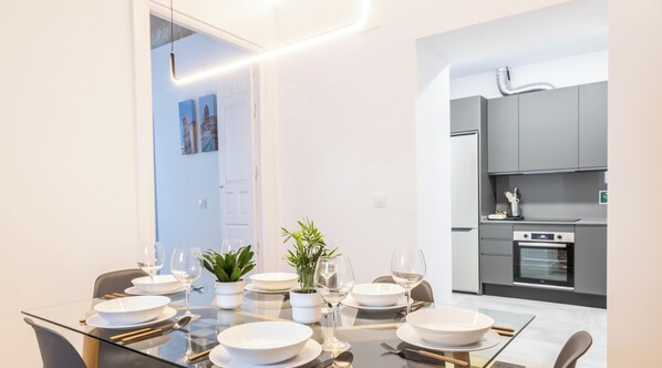 A modern dining area with a glass table, white plates, and wine glasses, leading to a kitchen with grey cabinets and a built-in oven.