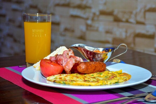 Un plato de desayuno con tomate, salchicha y tortilla, un vaso de zumo de naranja y una cuchara de plata sobre una mesa con un posavasos a rayas.