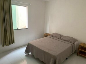 Une chambre avec un lit simple, une fenêtre avec des rideaux et une petite table de chevet en bois.