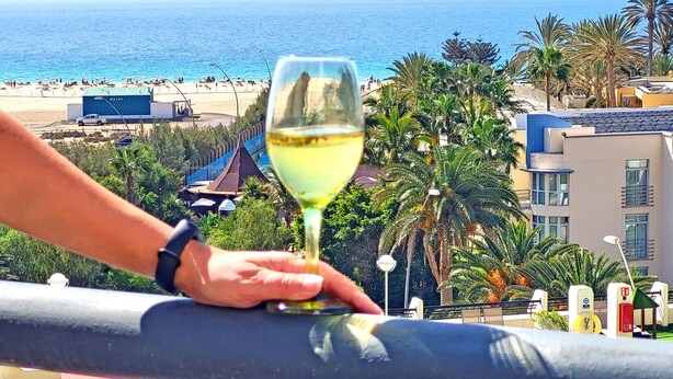 A balcony with a glass of white wine, overlooking a beach with palm trees and a clear blue sky. - Apartment BUENA VISTA (Palm Garden) Morro Jable, sea view, pool (Morro Jable)