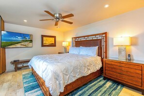 Une chambre avec un grand lit, une tête de lit en bois et un ventilateur de plafond. On y trouve une table de chevet avec une lampe, un tableau encadré au mur et un téléviseur fixé au mur.
