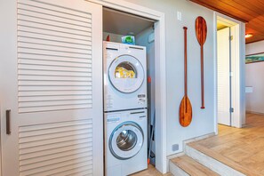 A laundry room with stacked washing machines, a wooden paddle, and a door leading to another room.