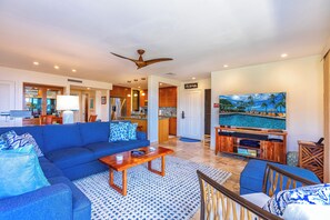 A modern living room with a blue sofa, wooden coffee table, and a TV on a wooden stand. - Ground Floor Luxury Condo with Stunning Panoramic Views – Wailea Elua #305 (Wailea, Maui)
