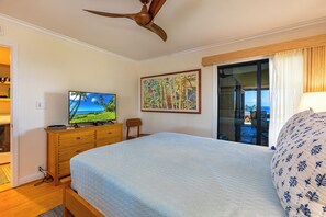 Un dormitorio con cama, televisor, cómoda de madera, ventilador de techo, un cuadro y vistas al exterior.