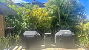 Un patio con dos asadores Weber, rodeado de exuberante vegetación tropical y suelo de baldosas.