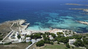 Une vue aérienne d’une zone côtière avec une plage, des eaux d’un bleu limpide et une petite ville avec des bâtiments et des voitures garées.