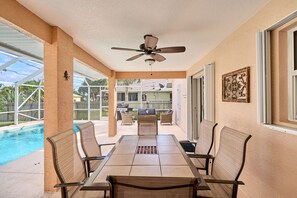 Un patio cubierto con una mesa y sillas, un ventilador de techo y vistas a una piscina y un edificio al fondo.