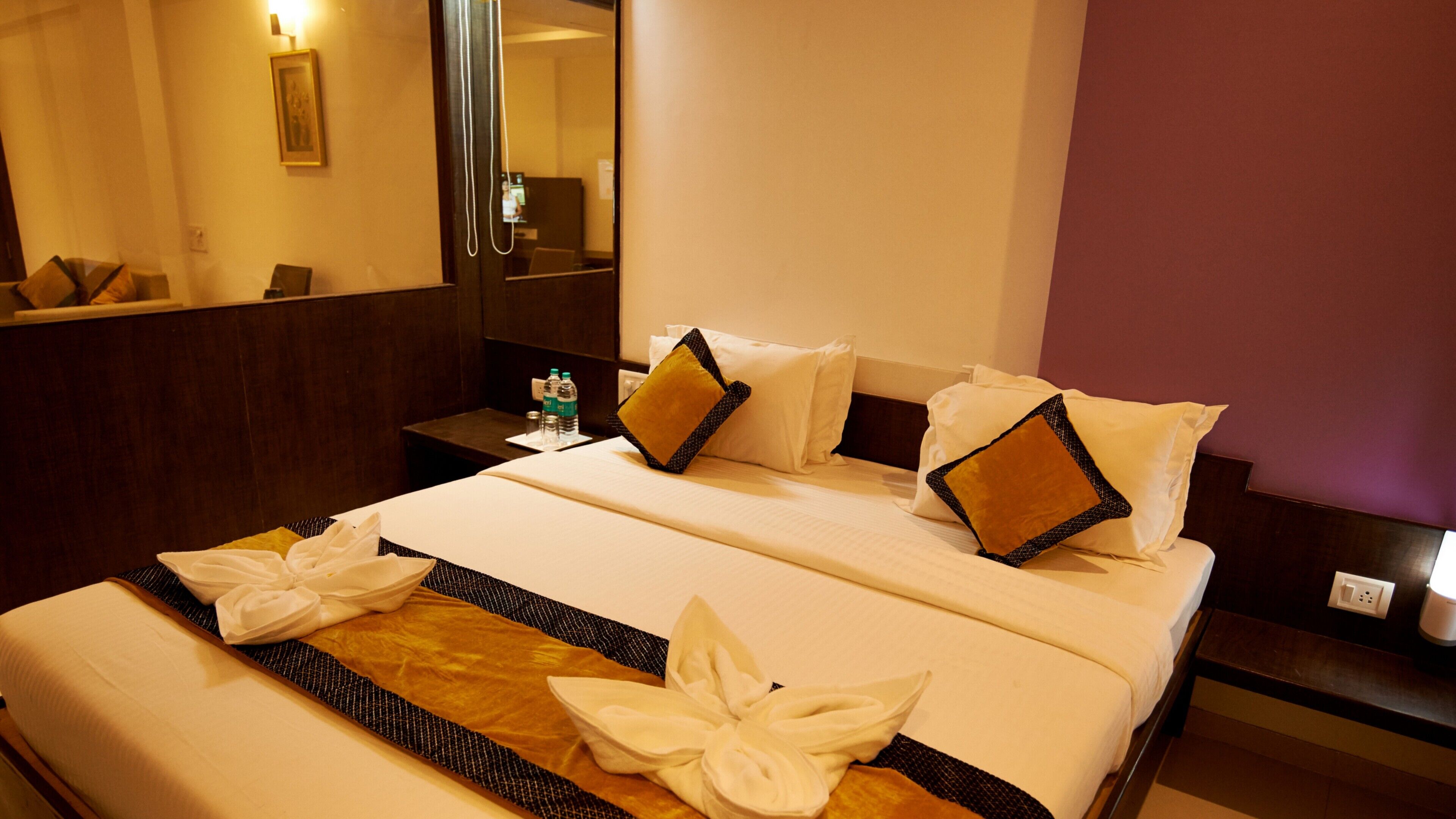 A hotel room with a neatly made bed, a bedside table with a water bottle, and a mirror on the wall.