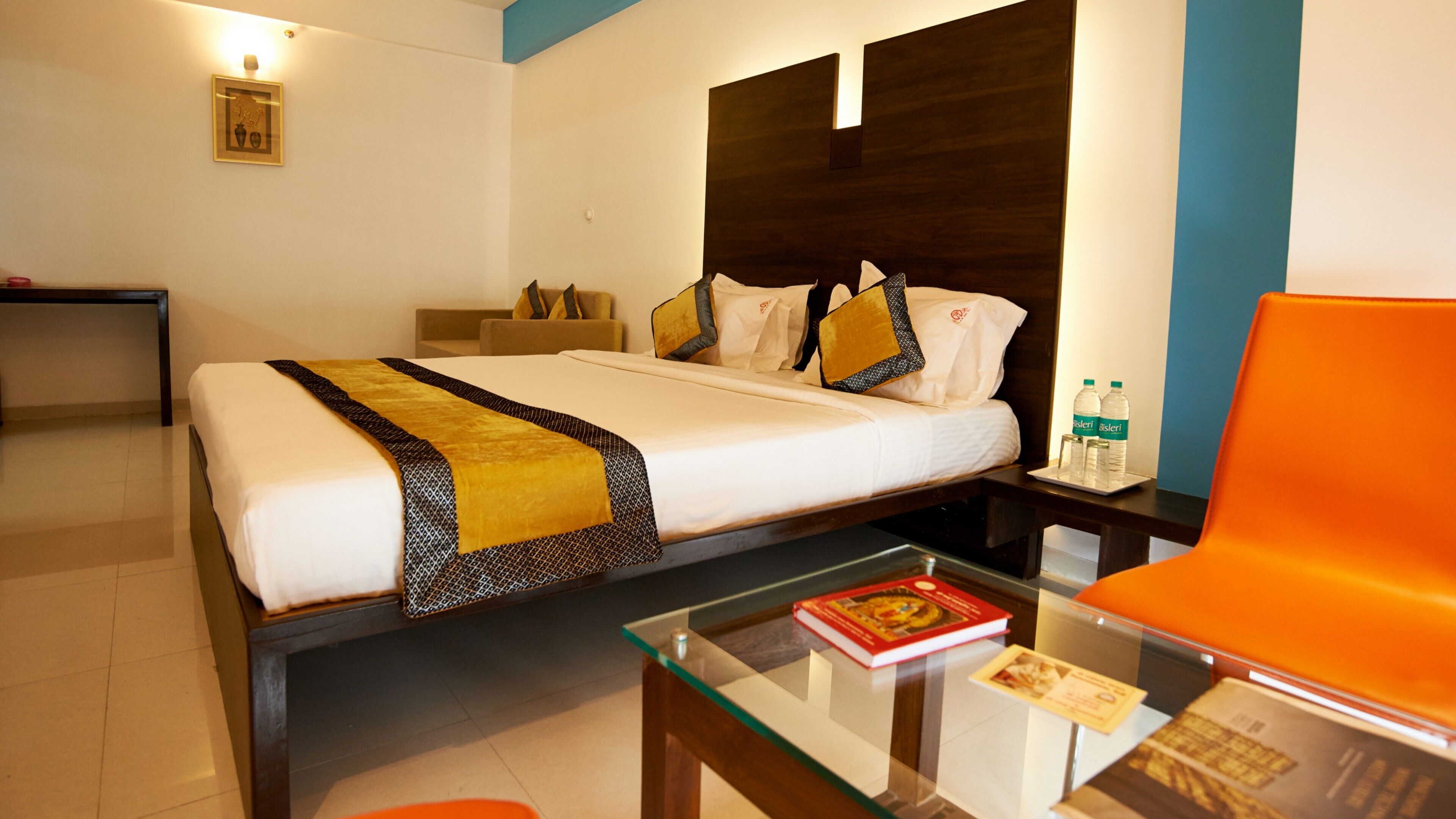 Una habitación de hotel con cama, una mesita de centro de vidrio con libros, una mesita de noche con botellas de agua y una silla.