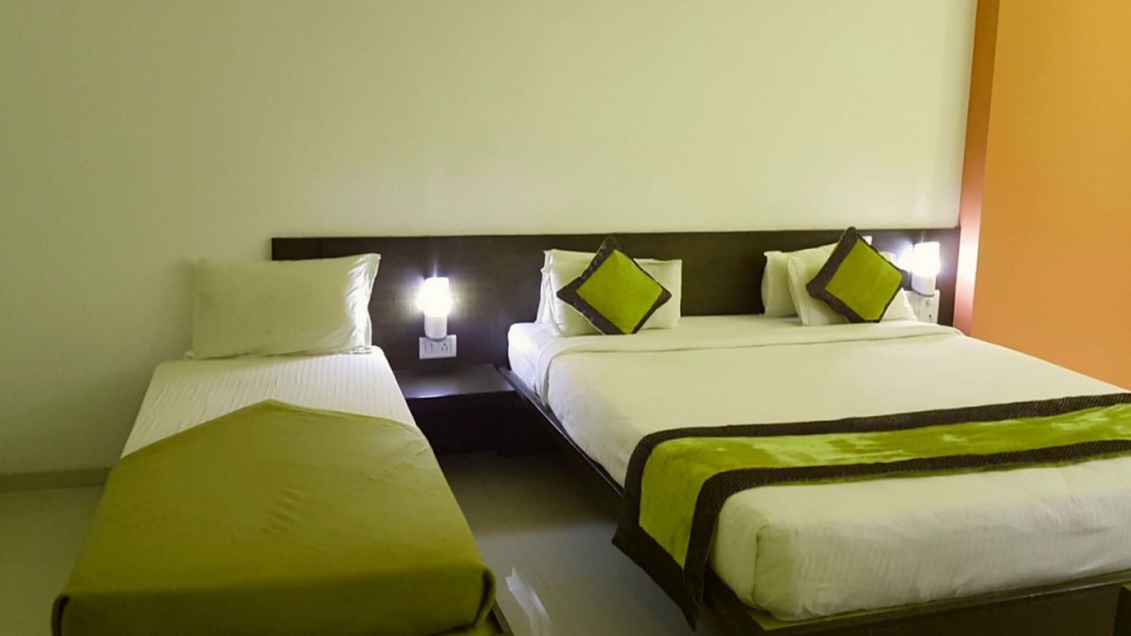 Dos camas individuales con ropa de cama blanca y detalles verdes, una mesita de noche con lámpara, y una pared lisa con interruptor de luz.