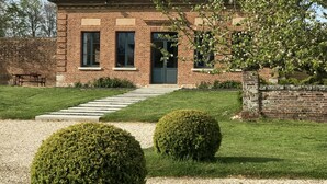A brick building with a red-tiled roof, a front door, and two windows. There is a gravel path leading to the entrance, and two well-maintained bushes in front. - The Château Orangerie (Hugleville-En-Caux)