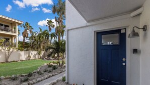 Eine blaue Tür mit der Nummer 114, eine weiße Wand und ein kleiner Garten mit Steinen und Pflanzen.
