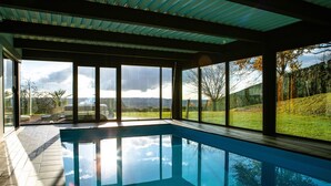 Ein moderner, überdachter Pool mit großen Fenstern und Blick auf eine grüne Landschaft und Bäume.