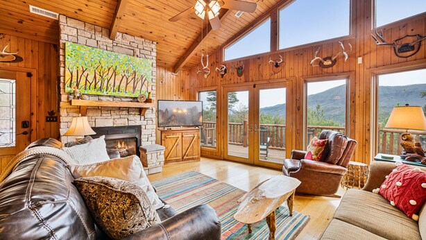 A cozy living room with a fireplace, leather sofas, a television, and a large window overlooking a mountainous landscape. - Amazing 5 br cabin-amazing mtn views & mins to NP (Sevierville)