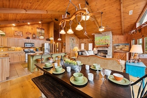 A wooden cabin interior with a dining area set for a meal, a kitchen with modern appliances, and a fireplace.
