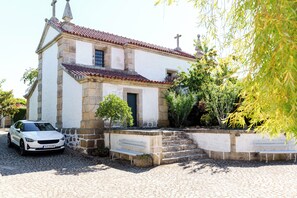 Traditional building with a red-tiled roof, featuring a cross, surrounded by lush greenery and a cobblestone street.