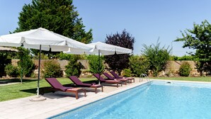 En swimmingpool med solstolar och parasoller, omgiven av grönska och en stenmur.