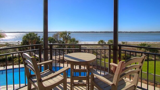 Un balcón con piscina, dos sillas de madera y una mesa con vista a una playa y al mar.