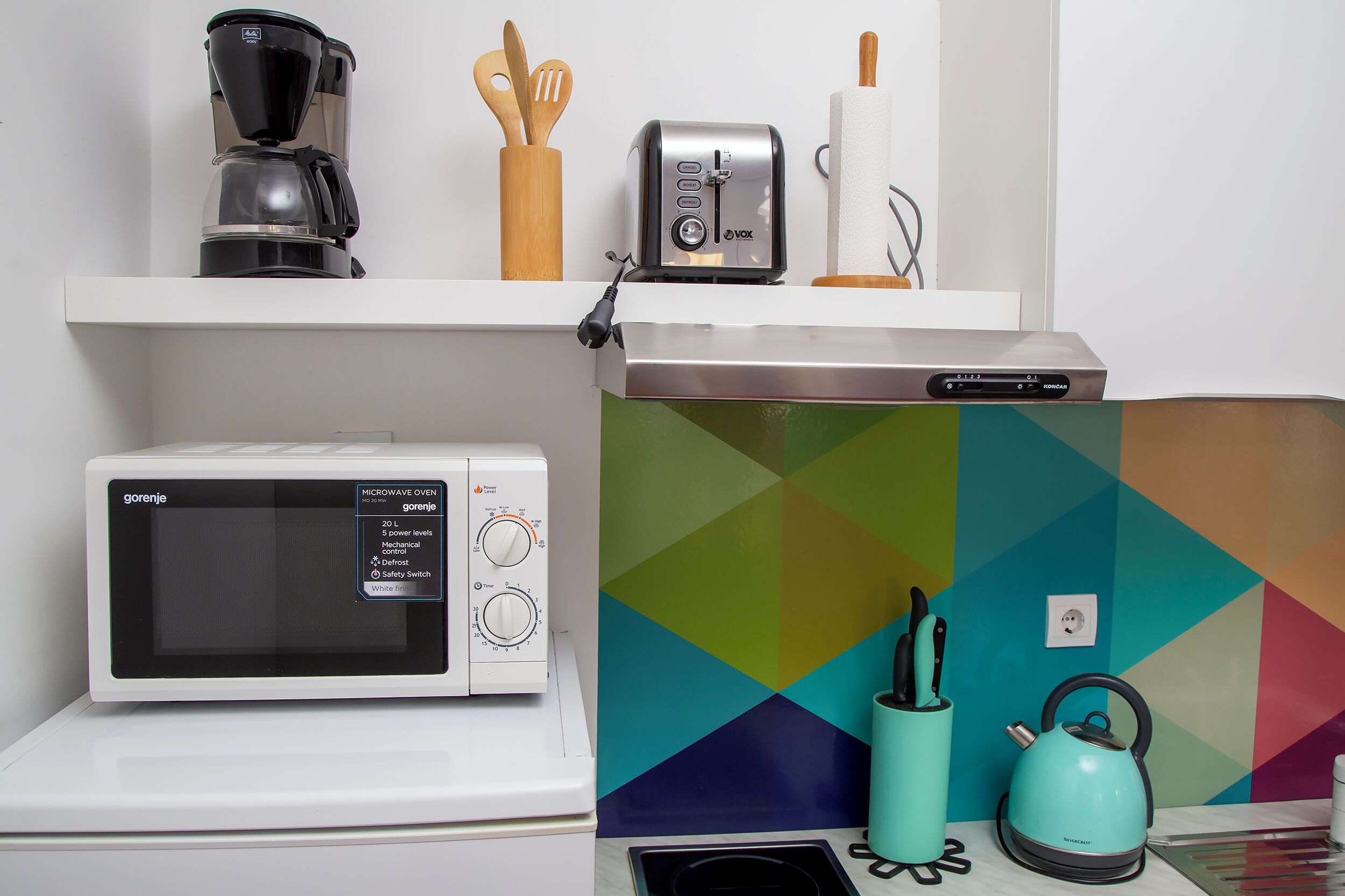A kitchen with a microwave, toaster, coffee maker, and a colorful geometric wall.