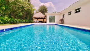 Ein klares, blaues Schwimmbecken mit einer gefliesten Kante, umgeben von üppigem Grün und einer strohgedeckten Struktur.