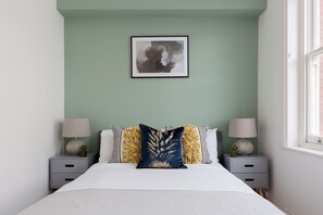 A neatly made bed with a white comforter and a decorative pillow, flanked by two bedside tables with lamps and small potted plants. A framed abstract artwork hangs above the bed. - Elegant Apartment near Spitalfields Market (London)