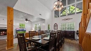 A dining area with a large wooden table, dark wooden chairs, and a chandelier. There is a fireplace with a stone mantle, and large windows with blinds.