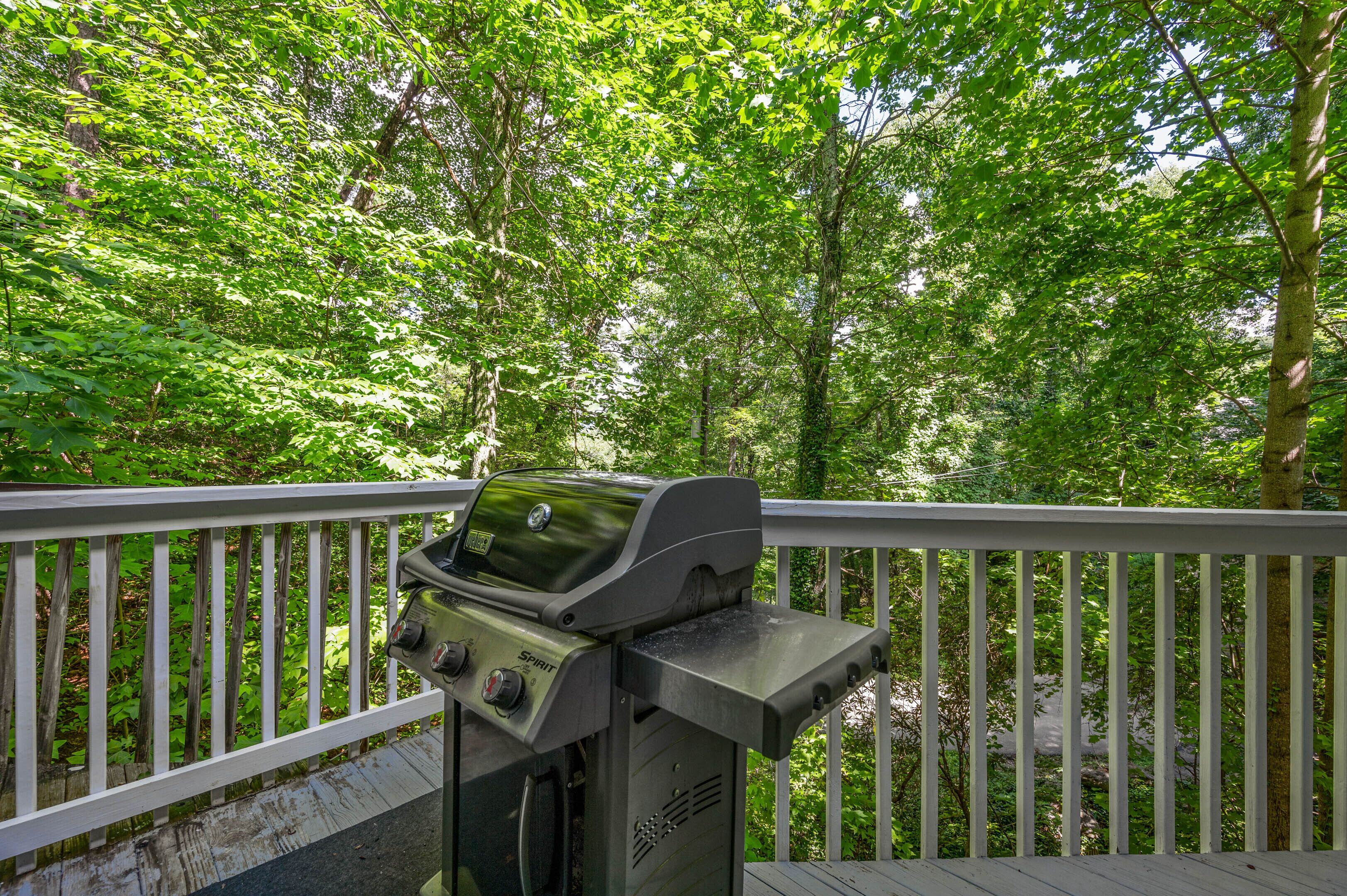 Een barbecue op een houten terras met een witte balustrade, omringd door weelderige groene bomen.