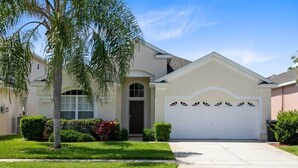 A single-story house with a white garage door, a front entrance with a brown door, and a small front yard with a palm tree and shrubs. - 4 Bedroom/3 Bath South Facing Pool/Spa Villa 4 Villa by RedAwning (Kissimmee)