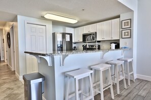 A modern kitchen with white cabinets, a stainless steel refrigerator, a microwave, and a built-in oven. There are four white bar stools and a trash can.