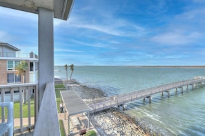 Vista costera con un muelle que se adentra en el agua, un edificio con balcón y cielo despejado.