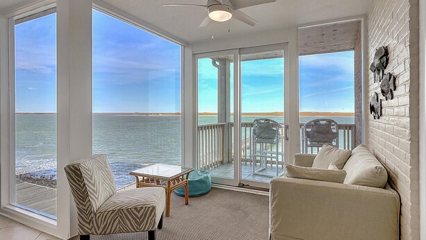 Une pièce lumineuse, baignée de soleil, avec une grande fenêtre donnant sur la mer, un ventilateur de plafond et un coin salon avec une table et des chaises.