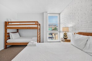 A room with a bunk bed, a single bed, a window with blinds, a lamp, and a white brick wall.