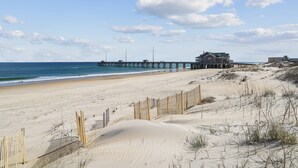 Ein Strand mit einer hölzernen Pier, die sich ins Meer erstreckt, einem sandigen Bereich mit Dünen und einem Gebäude mit einem großen Balkon mit Blick auf den Strand.