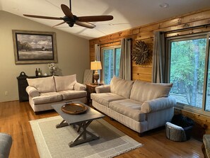 Ein Wohnzimmer mit zwei beigefarbenen Sofas, einem Holztisch, Holzboden, Deckenventilator, einem Fenster mit Vorhängen und einer Wanddekoration.