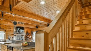 Interior de una cabaña de madera con cocina, chimenea de piedra y un amplio ventanal.