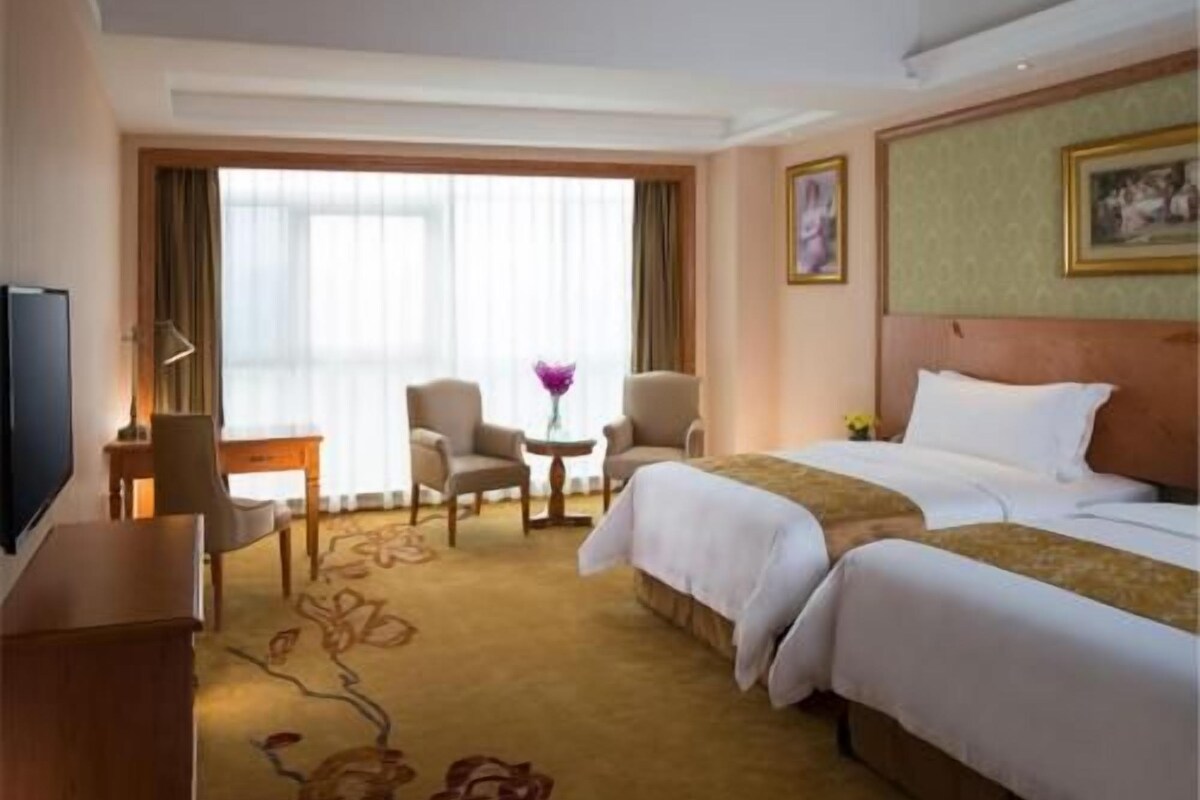 A hotel room with a bed, two armchairs, a small table, a TV, and a window with curtains.