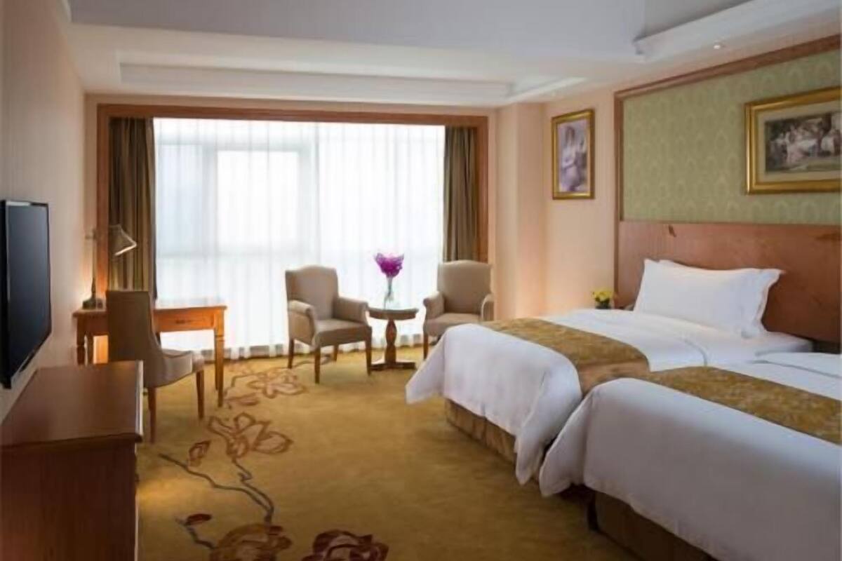 A hotel room with a bed, two armchairs, a small table, a TV, and a window with curtains.