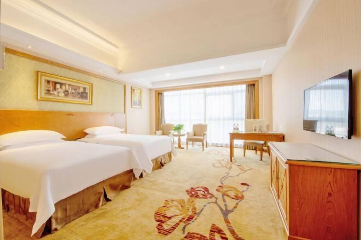 A hotel room with two beds, a desk, a television, and a large window.