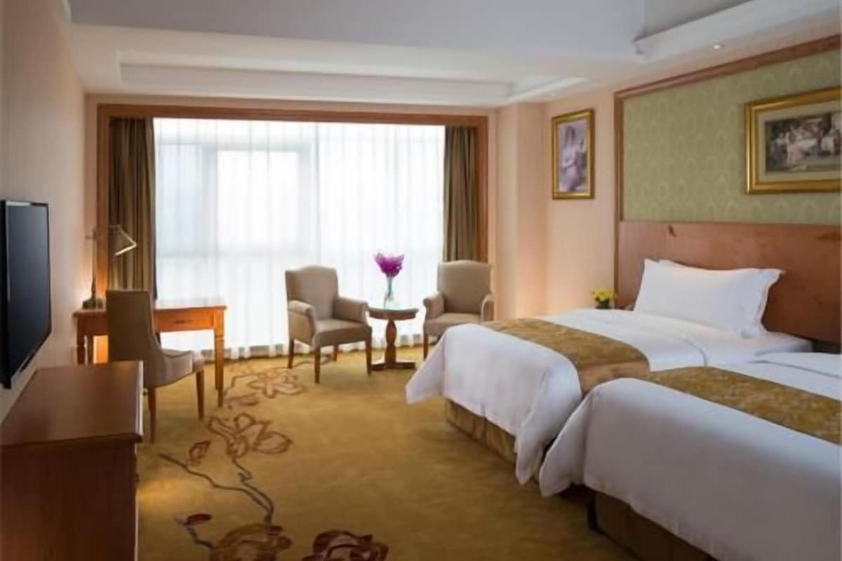 A hotel room with a bed, two armchairs, a small table, a TV, and a window with curtains.
