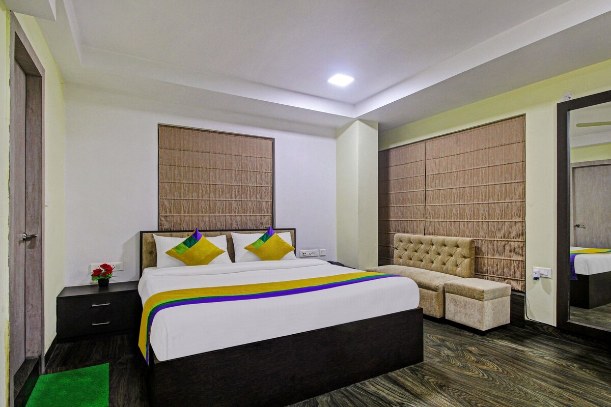 A hotel room with a large bed, a chest of drawers, a bedside table, a mirror, and a window with a view.
