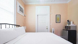Une chambre avec un lit, une porte blanche, un climatiseur fixé au mur, un petit réfrigérateur et un tableau accroché au mur.
