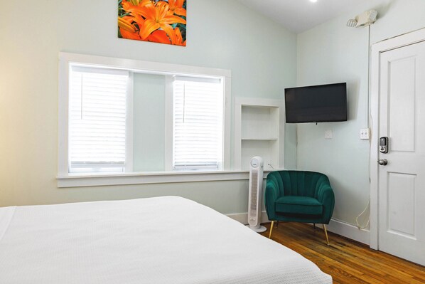 Una habitación con cama, ventana con persianas, una silla color turquesa, un televisor y una puerta.