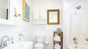 Un baño con lavamanos, inodoro y bañera blancos. Hay armarios blancos sobre el inodoro y un estante con toallas.