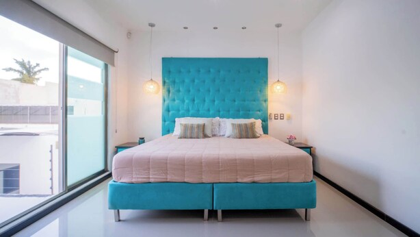 A modern bedroom with a large bed, a teal headboard, and a large window offering a view of the outside.