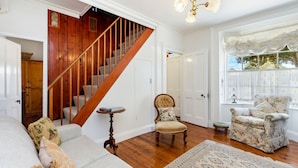 A spacious living room with a wooden staircase, a floral armchair, and a sofa with patterned cushions. - Norina Cottage - circa 1858 (Port Fairy)