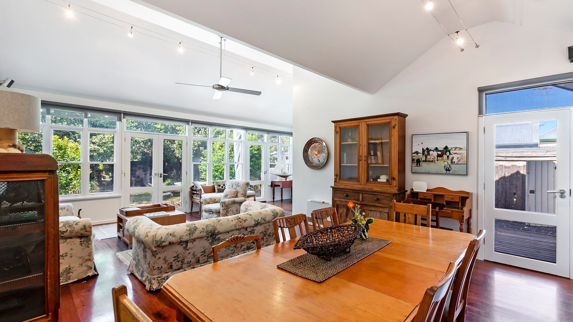 A spacious dining area with a wooden table, floral sofa, and a large window offering a view of greenery.