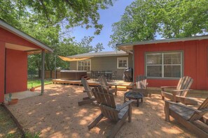 A covered outdoor patio with wooden chairs and a fire pit, surrounded by a red building with a large window and a door. - Modern Comfort-Hot tub, Firepit, Mins to Downtown! (Fredericksburg)