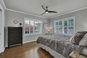 A bedroom with a bed, dresser, ceiling fan, and large windows.