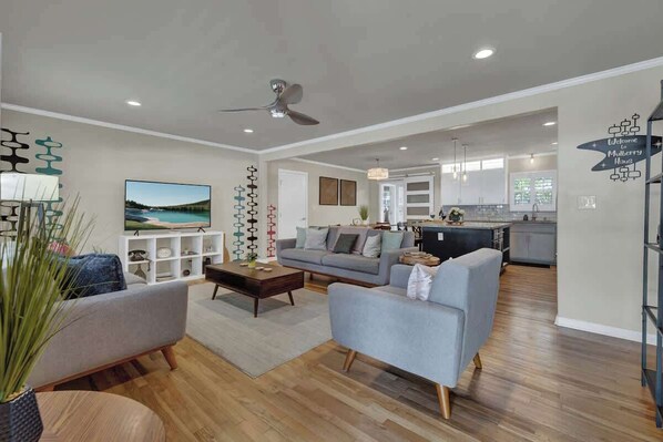 A spacious living room with a grey sofa, a coffee table, and a TV. There is a kitchen in the background with modern appliances and a sign that reads "Welcome to Mulberry House."